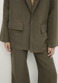 Olijfgroene blazer met twee voorkeurszakken en een relaxte pasvorm, gecombineerd met bijpassende wijde broek. Glad linnen-achtig textuur.