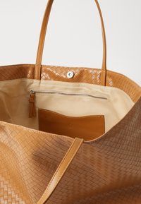 Tan geweven leren tote bag met twee handvaten, met een ruime binnenvoering en een ritsvak tegen een beige voering.