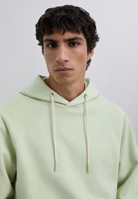 Young man with short curly dark hair wearing a light green hoodie, looking straight ahead with a neutral expression against a plain background.
