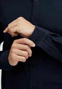 Hombre abrochando un botón negro en la manga larga de una camisa azul marino, mostrando un primer plano de las manos y los detalles de la camisa.