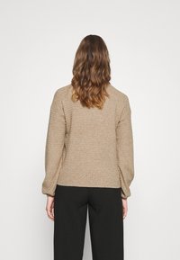 Beżowy sweter z dzianiny z teksturowanym heksagonalnym wzorem, z długimi rękawami i luźnym krojem, w połączeniu z ciemnymi spodniami.