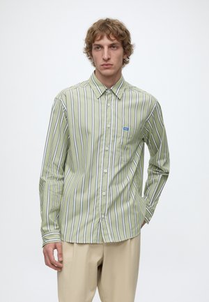 Jeune homme aux cheveux bouclés portant une chemise à rayures verticales vertes et blanches et un pantalon beige, debout avec une main dans la poche.