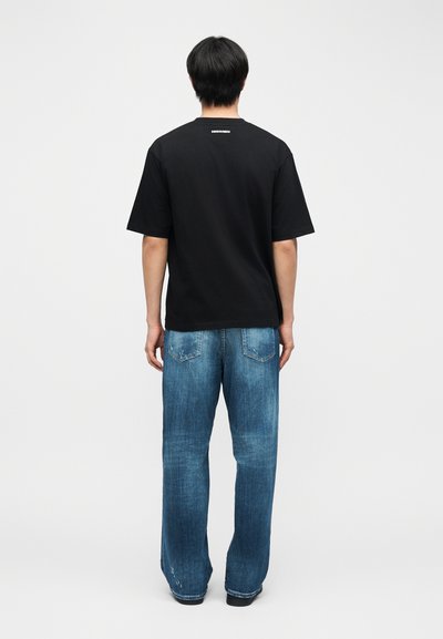 Άτομο που στέκεται με την πλάτη γυρισμένη, φορώντας μαύρο κοντομάνικο μπλουζάκι και άνετο μπλε τζιν, μπροστά σε απλή φόντο.