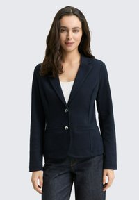 Navy blazer made of textured fabric, features a notched lapel, two buttons, and side pockets, paired with dark jeans.