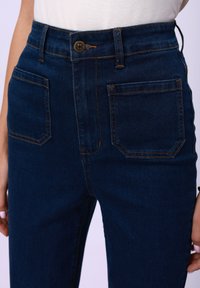 Hochgeschnittene dunkelblaue Jeans aus Denim mit braunen Nähten, ausgestattet mit zwei Fronttaschen und zwei Gesäßtaschen, Knopfverschluss an der Taille.