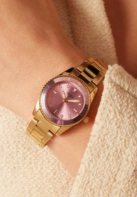 Gouden polshorloge met een roze wijzerplaat, gemarkeerde uur indicatoren en een bijpassende bezel. Voorzien van een metalen band en een gestructureerde stoffen achtergrond.