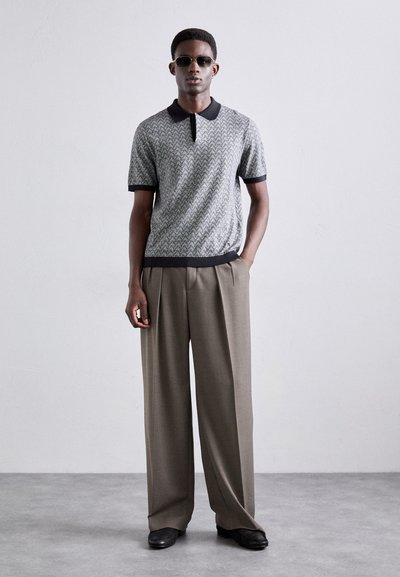Polo shirt à motifs gris avec col noir, associé à un pantalon large beige. Le mannequin porte des lunettes de soleil sombres. Fond minimaliste.