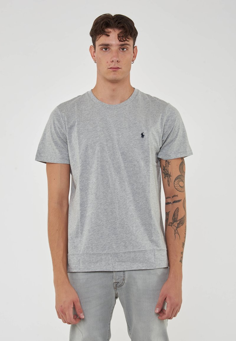 T-shirt in cotone grigio con scollo rotondo, maniche corte e un logo blu navy discreto sul petto. Orlo dritto, abbinato a jeans grigi chiari.