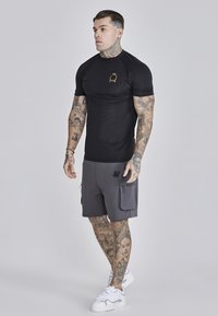 Schwarz enges T-Shirt mit einem goldenen Logo, kombiniert mit grauen Sportshorts mit zwei Seiten Taschen. Weiße Sneakers runden den Look ab.