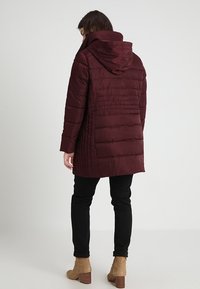 Cappotto imbottito bordeaux con cappuccio, caratterizzato da cuciture orizzontali, maniche lunghe e tasche laterali. Indossato sopra pantaloni neri e stivaletti alla caviglia.