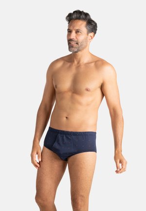 Homme torse nu aux cheveux foncés portant un slip bleu marine avec de petits motifs rouges, debout devant un fond clair uni, regardant vers la droite.