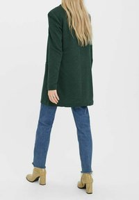 Grön lång kavaj i mjukt material, med en enkel design. Bärs över blå denimjeans och guldklackade skor.