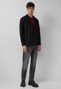 Schwarzes Langarmhemd mit Knöpfen und Brusttaschen, über einem roten T-Shirt getragen. Kombiniert mit grauen Jeans und schwarzen Knöchelschuhen.