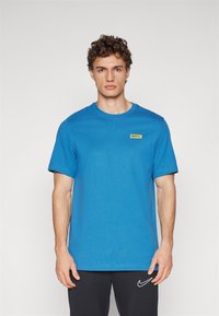 Camiseta de algodón azul con cuello redondo y mangas cortas. Presenta un pequeño logo amarillo de Nike F.C. en el lado izquierdo del pecho.