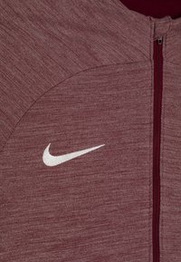 Bordeauxkleurige zip-upjas met een gemêleerd textuur en een contrasterend wit Nike-logo op de borst; beschikt over een hoge kraag en een ritssluiting.
