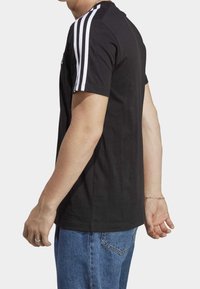 T-shirt in cotone nero con design a tre righe bianche sulle spalle, collo a giro, maniche corte e vestibilità rilassata. Indossato con jeans in denim blu.