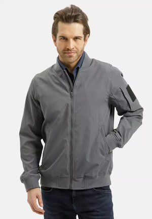 Mężczyzna w szarej kurtce zapinanej na zamek, z prążkowanymi mankietami i kołnierzem, z ręką w kieszeni, stojący na białym tle.