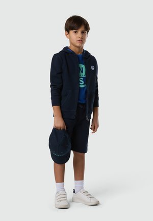 Niño con sudadera con capucha azul marino, camiseta azul, pantalones cortos azul marino, zapatillas blancas, calcetines blancos, sosteniendo una gorra de béisbol azul marino sobre un fondo liso.
