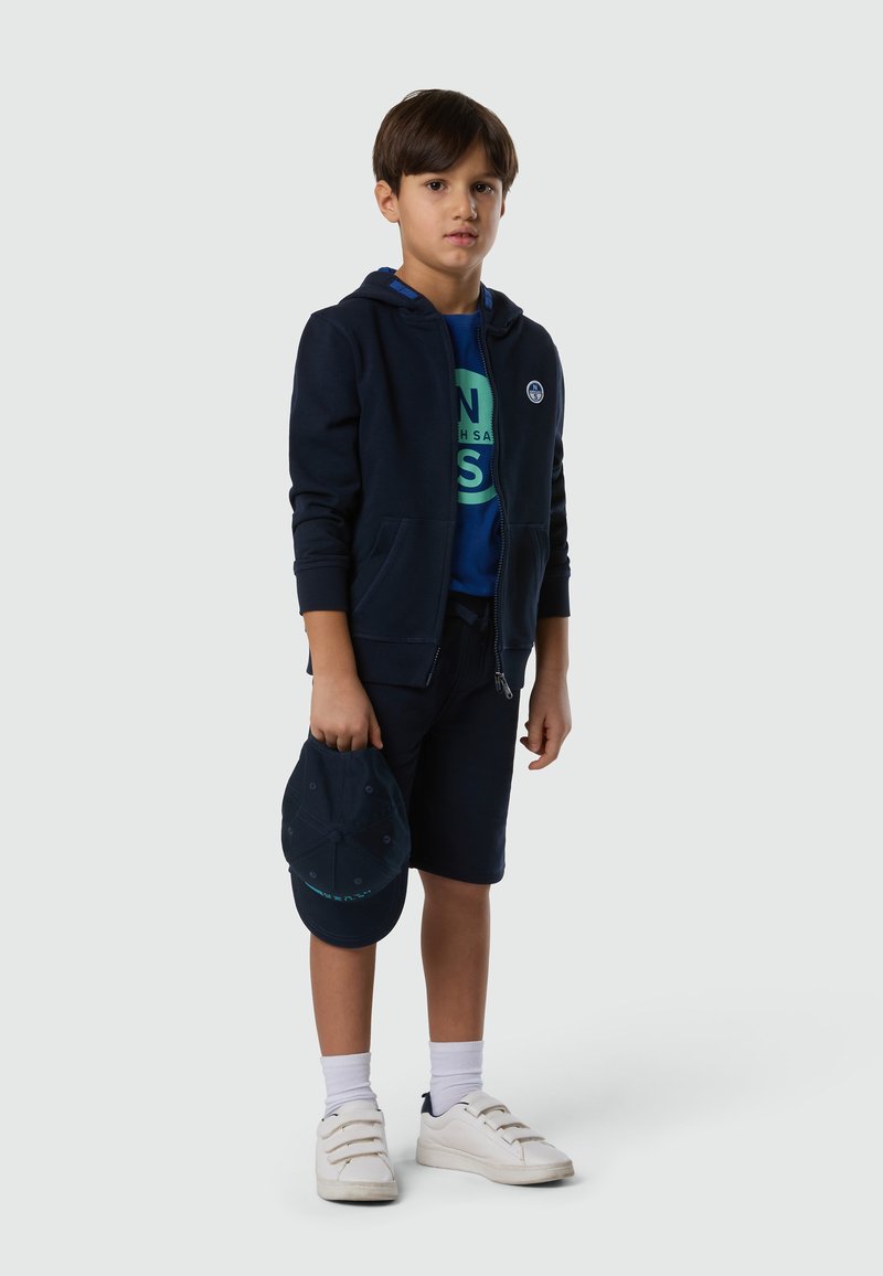 Ragazzo con felpa con cappuccio blu navy, t-shirt blu, pantaloncini blu navy, sneakers bianche, calzini bianchi, che tiene un cappellino da baseball blu navy su uno sfondo semplice.