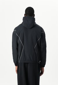 Veste noire légère avec capuche, dotée de coutures blanches, de poignets élastiques et d'un ourlet resserré. Design simple, texture douce.