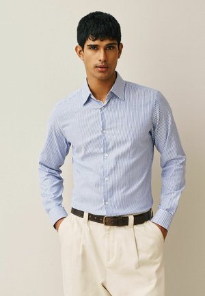 Mann trägt ein blau-weiß vertikal gestreiftes Hemd mit Knöpfen, in cremefarbene Hosen gesteckt, mit einem dunkelbraunen Gürtel, Hände in den Taschen.