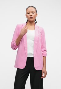 Ružový blazer so štíhlym strihom, s klopovým golierom a mierne prehnutými rukávmi, skombinovaný s bielym topom a čiernymi nohavicami. Hladká textúra látky.