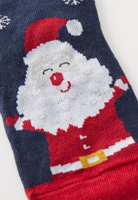 Marinblå strumpa med design av en röd julsniske med vit skägg, röd näsa och guldbälte. Snöflingemönster ovanför figuren.