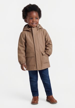 Smilende barn iført en brun hættejakke, blå jeans og brune støvler, stående med den ene hånd i jakke-lommen.