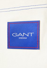 Weißer Stoff mit blauen und roten Details; blauer rechteckigerPatch mit "GANT" und "SPORTSWEAR" in weißem, zentriertem Text.