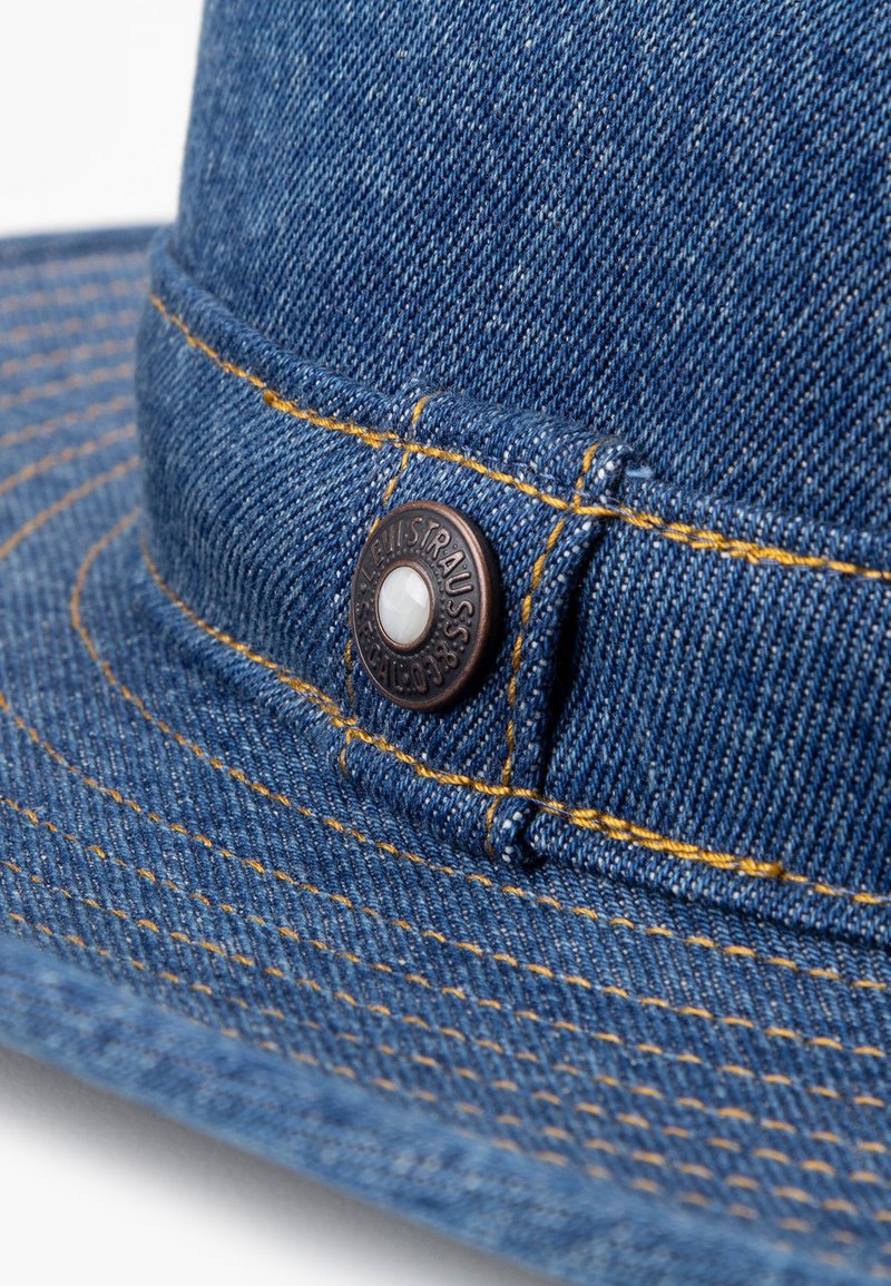 Cappello di jeans con visiera larga, cuciture gialle a contrasto e un bottone con logo circolare marrone scuro con un dettaglio centrale bianco.