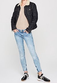 Personne portant un jean skinny bleu clair, une veste en denim noir avec un col beige, une chemise rayée beige et des baskets noires avec des lacets blancs.