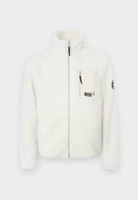 Veste en polaire blanche avec un col montant, une fermeture éclair à l'avant et une poche poitrine. Elle présente une texture douce et un patch logo sur la manche.