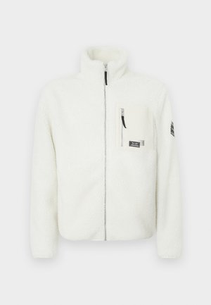 Chaqueta de forro polar blanca con cuello alto, cierre de cremallera en la parte delantera y un bolsillo en el pecho. Presenta una textura suave y un parche con el logo en la manga.