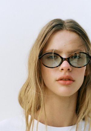 Tânără cu părul lung și blond purtând ochelari rotunzi supradimensionați cu ramă de tortoiseshell, expresie neutră, pe un fundal simplu și luminos.