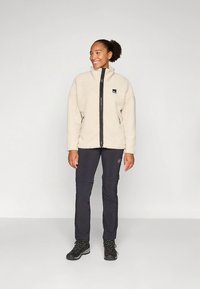 Beige fleecejakke med glidelås foran, svarte detaljer og en liten logo på brystet, kombinert med mørke bukser og svarte tursko.