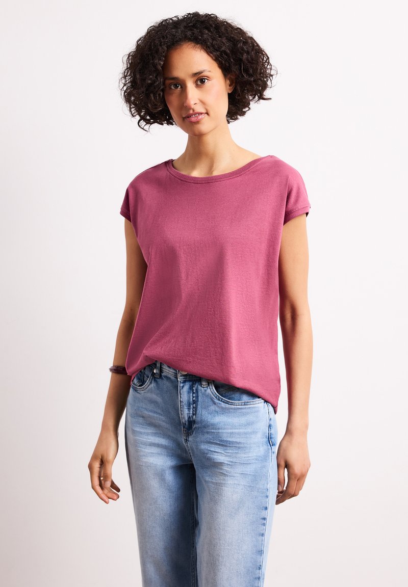 Donna con capelli corti e ricci che indossa una maglietta rosa a maniche corte e jeans azzurri, in piedi davanti a uno sfondo bianco semplice.