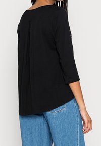 Sort langærmet top med rund halsudskæring og plissering bagpå; glat tekstur; parret med lyseblå denimjeans.