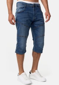 Pantaloni capri din denim, de culoare albastră, cu un croi ajustat, multiple linii de cusătură și o margine răsucită. Purtați cu adidași albi pentru un aspect casual.