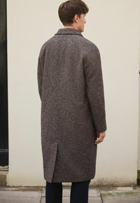 Lang grå uldfrakke med en meleret mønster, slagkrave og en enkelt slids bagpå; parret med mørke jeans.