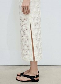Weißes, gesticktes Kleid mit floralem Muster und seitlichem Schlitz; kombiniert mit schwarzen flachen Sandalen mit Schnallen- und Riemen-Design.