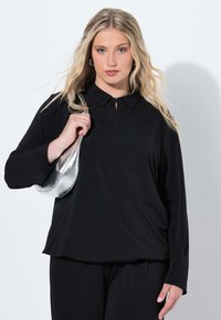 Top nero a maniche lunghe con colletto e dettaglio a chiave sul fronte. La modella tiene una borsa metallica argentata. Tessuto liscio con vestibilità rilassata.