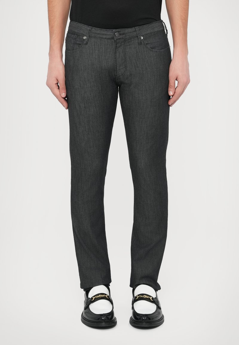 Temno sivi slim-fit jeans iz blaga z rahlo teksturo. Imajo pet žepov in kovinske detajle, kombinirani s črnimi loaferji in belimi nogavicami.