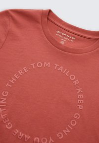 Rotes Baumwoll-T-Shirt mit Rundhalsausschnitt, das erhobene Textgrafiken in kreisförmiger Anordnung zeigt. Das Etikett weist auf die körpernahe Passform in Größe S/140 hin.