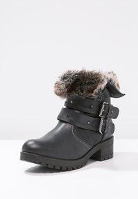 Černá kotníková bota s masivním podpatkem, dvěma přezkami a kožešinovým lemováním kolem horního okraje na bílém pozadí.