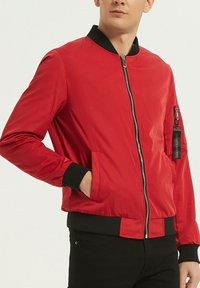 Blouson aviateur rouge en tissu lisse et brillant. Col et poignets en ribbed noir. Doté d'une fermeture éclair et d'une poche latérale avec zip.