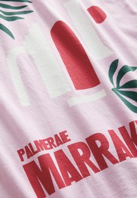 Heledast roosa puuvillane t-särk, millel on suured punased ja valged tähestikud "PALMERAIE MARRAKECH" ning rohelised palmi lehe graafilised kujutised.