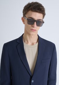 Blazer navy in un tessuto liscio con un solo bottone, abbinato a una camicia beige. Occhiali da sole scuri con montatura rettangolare.