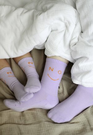 Pieds d’un adulte et d’un enfant portant des chaussettes lavande assorties avec des visages souriants orange, partiellement couverts par une couverture blanche sur un lit beige.