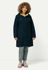 Cappotto impermeabile navy con cappuccio, foderato internamente di un vivido verde, tasche frontali e una vestibilità comoda. Abbinato a jeans azzurri e stivali rosa.