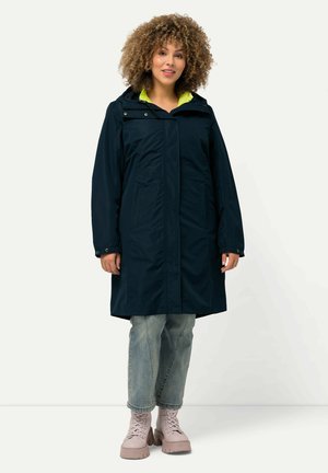 Navy waterproof coat with a hood, a bright green inner lining, front pockets, and a relaxed fit. Paired with light blue jeans and pink boots.